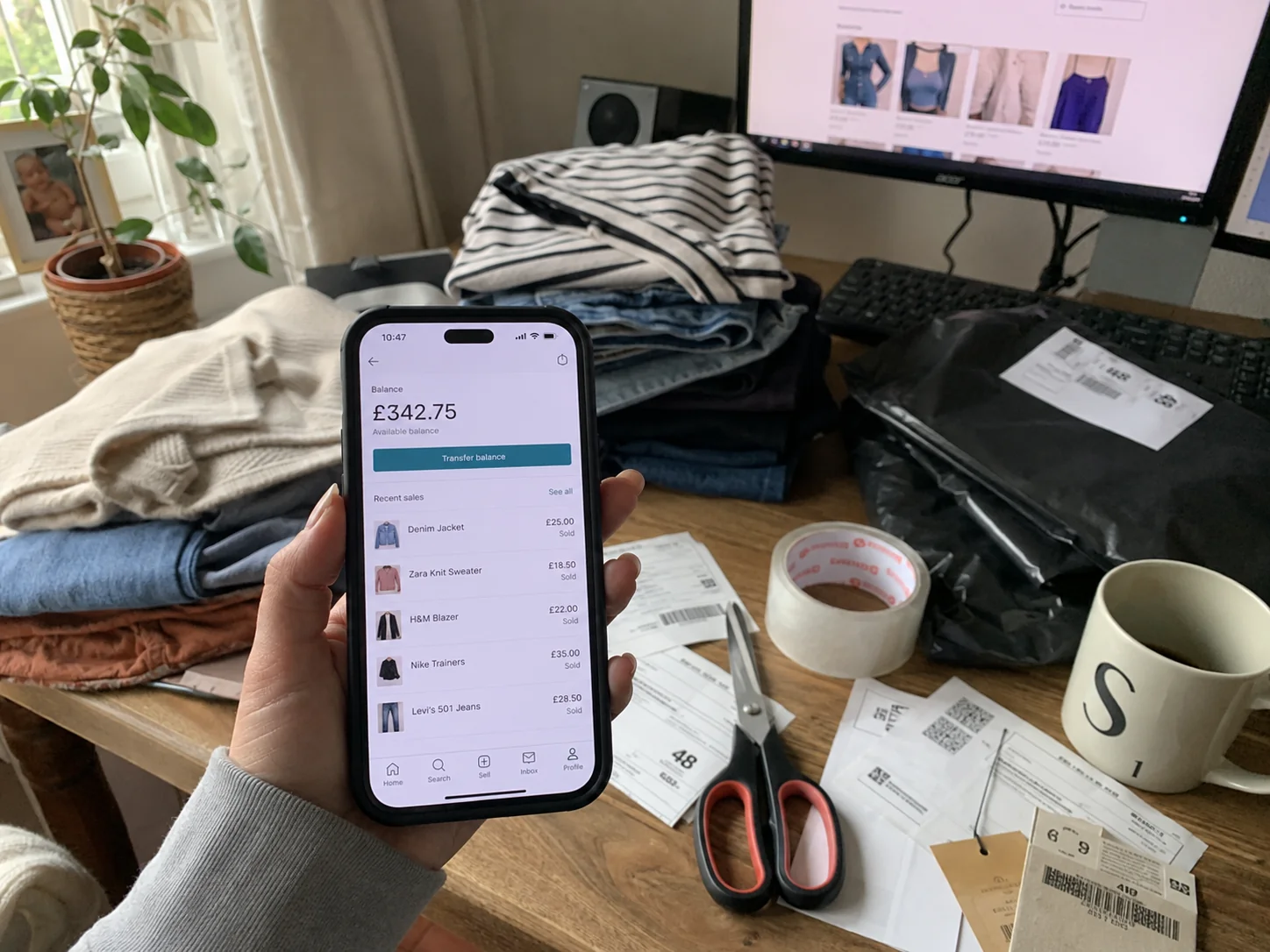 A hand holding a phone showing a £342.75 selling balance, over a desk of folded clothes and packed parcels